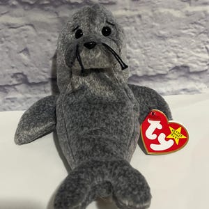 May include: A gray plush seal toy with black eyes and whiskers. The seal has a red heart-shaped tag with white text and a yellow heart-shaped tag with black text. The toy is on a white surface.