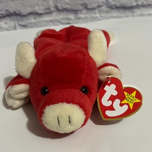 May include: A red plush toy bull with cream-colored ears, snout, and hooves. The toy has black eyes and a tag that reads "BEANIE ORIGINAL BABY". The bull is lying down on a white surface.