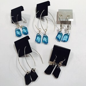 May include: Three pairs of earrings displayed on black card stock. Two pairs feature blue crystal teardrop shapes on silver hoops. One pair features black crystal teardrop shapes on silver hoops.