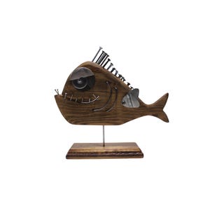 Könnte beinhalten: Eine dekorative Fischskulptur aus dunkel gebeiztem Holz mit einer großen Metallscheibe als Auge, einer Reihe von Metallnägeln auf der Oberseite als Rückenflosse, einer seitlichen Metallflosse und einem gebogenen Metalldraht, der einen Mund bildet.