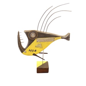 Könnte beinhalten: Eine Fischskulptur aus verschiedenen Metall- und Holzkomponenten. Der Körper ist gelb und dunkelbraun mit einer strukturierten Metallseitenwand, einem kreisförmigen Auge, scharfen Metallspitzen als Zähne und langen, gebogenen Drahtflossen.