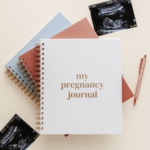 May include: A white pregnancy journal with gold text, "my pregnancy journal", and a spiral binding. It is next to notebooks in pink and blue, a glitter pen, and sonogram images. The notebooks are in cm.