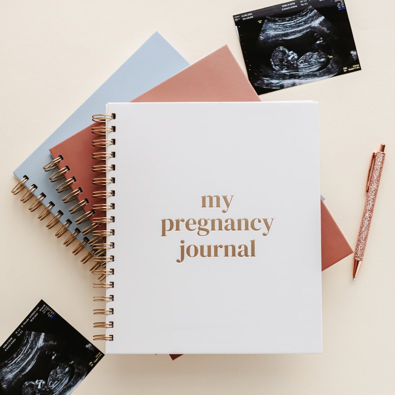 May include: A white pregnancy journal with gold text, "my pregnancy journal", and a spiral binding. It is next to notebooks in pink and blue, a glitter pen, and sonogram images. The notebooks are in cm.