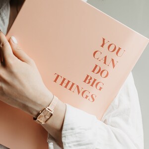 May include: A peach-colored notebook with the words "YOU CAN DO BIG THINGS" printed in a warm, reddish-brown font. A gold watch with a rectangular face is visible.