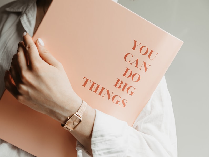 May include: A peach-colored notebook with the words "YOU CAN DO BIG THINGS" printed in a warm, reddish-brown font. A gold watch with a rectangular face is visible.