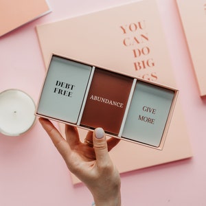 May include: A rectangular box with three cards, each with a word: "DEBT FREE", "ABUNDANCE", and "GIVE MORE". The box is held against a pink background with a candle and a notebook. The words "YOU CAN DO BIG THINGS" are on a notebook.