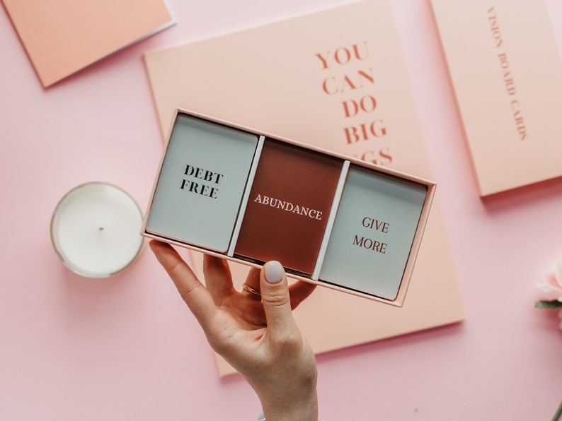 May include: A rectangular box with three cards, each with a word: "DEBT FREE", "ABUNDANCE", and "GIVE MORE". The box is held against a pink background with a candle and a notebook. The words "YOU CAN DO BIG THINGS" are on a notebook.