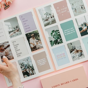 May include: Open vision board with inspirational cards featuring quotes like "Your potential to succeed is infinite" and "It's a good day to have a good day." The board is displayed on a pink surface with a box of cards.