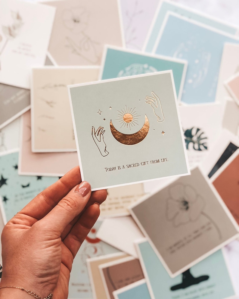 May include: A light blue card with a gold foil design of a crescent moon, sun, and two hands reaching up. The text on the card reads "Today is a sacred gift from life."
