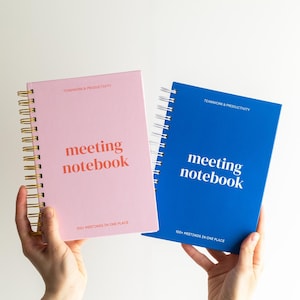 May include: A pink spiral-bound notebook with gold wire and a blue spiral-bound notebook with silver wire. Both notebooks feature the text "TEAMWORK & PRODUCTIVITY" at the top and "meeting notebook" in the center, with "100+ MEETINGS IN ONE PLACE" below.