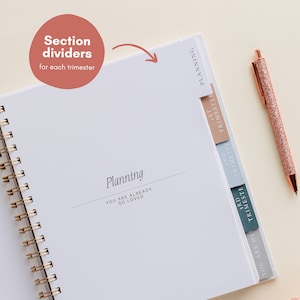 May include: A white spiral-bound planner with section dividers marked "1st Trimester", "2nd Trimester", and "3rd Trimester". The cover reads "Planning" and "YOU ARE ALREADY SO LOVED". A rose gold pen with glitter detail lies beside the open planner.