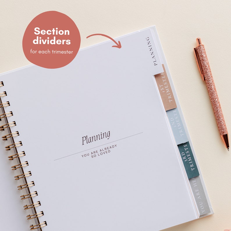 May include: A white spiral-bound planner with section dividers marked "1st Trimester", "2nd Trimester", and "3rd Trimester". The cover reads "Planning" and "YOU ARE ALREADY SO LOVED". A rose gold pen with glitter detail lies beside the open planner.