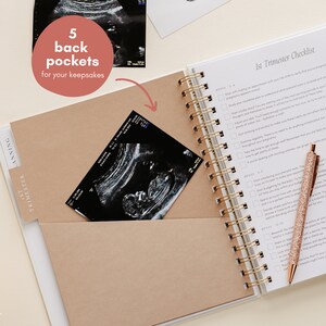 May include: A white pregnancy planner with a gold spiral binding and a rose gold pen. The planner features a pocket containing an ultrasound scan. Text on the planner reads "1st Trimester Checklist" and "5 back pockets for your keepsakes."