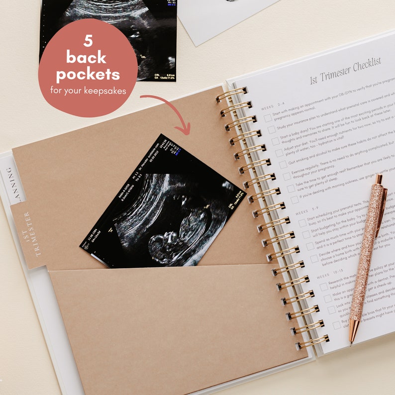 May include: A white pregnancy planner with a gold spiral binding and a rose gold pen. The planner features a pocket containing an ultrasound scan. Text on the planner reads "1st Trimester Checklist" and "5 back pockets for your keepsakes."