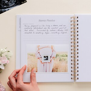 May include: A white spiral-bound journal with gold detailing, open to a page titled "Maternity Photoshoot." The page features handwritten text and a photograph of a pregnant person holding an ultrasound image. An ultrasound scan is visible.