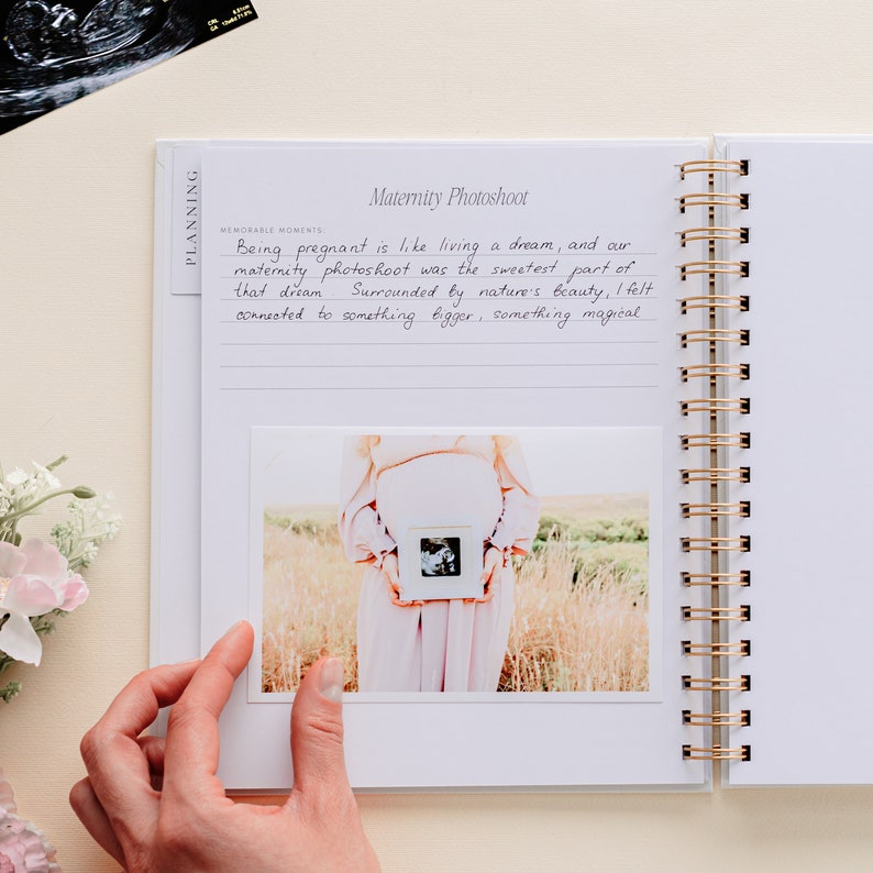 May include: A white spiral-bound journal with gold detailing, open to a page titled "Maternity Photoshoot." The page features handwritten text and a photograph of a pregnant person holding an ultrasound image. An ultrasound scan is visible.