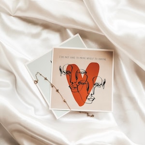 May include: A white card with the text "I do not have to prove myself to anyone" and a red heart with three faces drawn in black lines. A gold chain necklace with small charms is on top of the card.