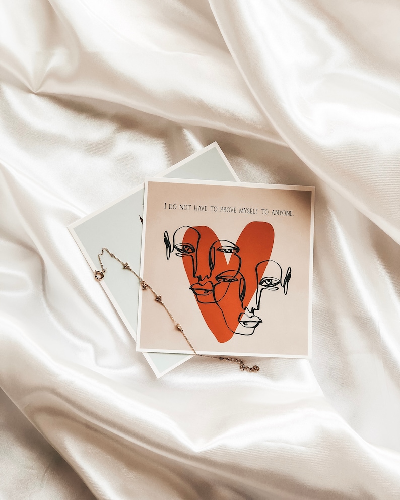 May include: A white card with the text "I do not have to prove myself to anyone" and a red heart with three faces drawn in black lines. A gold chain necklace with small charms is on top of the card.