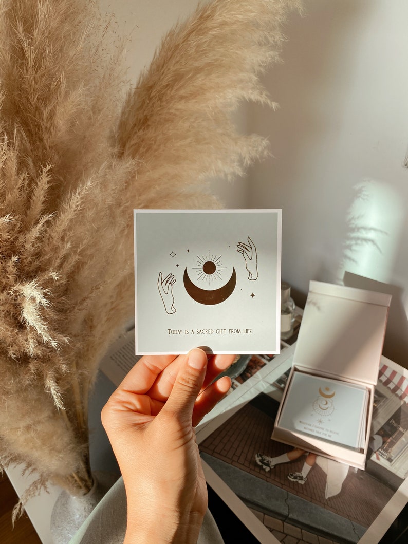 May include: A light blue card with a gold illustration of a sun, moon, and two hands reaching up. The text on the card reads "Today is a sacred gift from life."