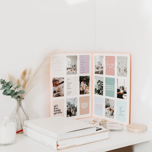 May include: Aesthetic desk setup featuring a journal with inspirational quotes, a glass vase with eucalyptus and pampas grass, a candle, and a stack of books. The journal is open, displaying colorful cards with motivational phrases.