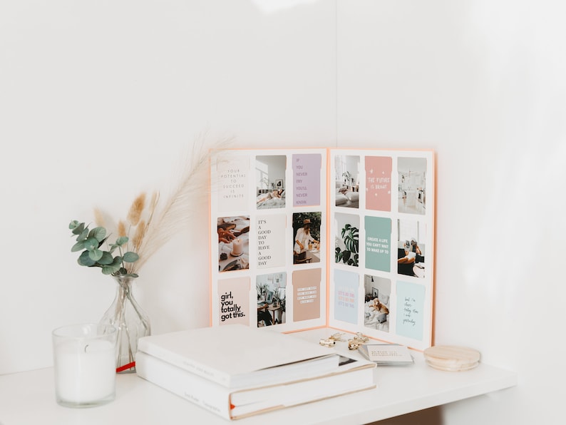 May include: Aesthetic desk setup featuring a journal with inspirational quotes, a glass vase with eucalyptus and pampas grass, a candle, and a stack of books. The journal is open, displaying colorful cards with motivational phrases.