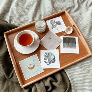 May include: A wooden tray with a cup of tea, a candle, and several cards with inspirational quotes and illustrations of plants and celestial bodies. The tray is on a green and white striped blanket.