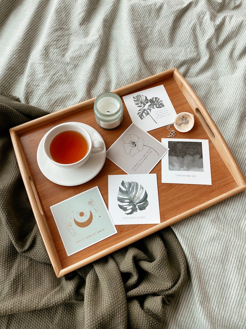 May include: A wooden tray with a cup of tea, a candle, and several cards with inspirational quotes and illustrations of plants and celestial bodies. The tray is on a green and white striped blanket.