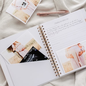 May include: A white spiral-bound journal with gold detailing, open to a page with handwritten text and a photo. Several photos and a pen with a glittery rose gold finish are also visible. The journal has pockets holding photos and an ultrasound image.