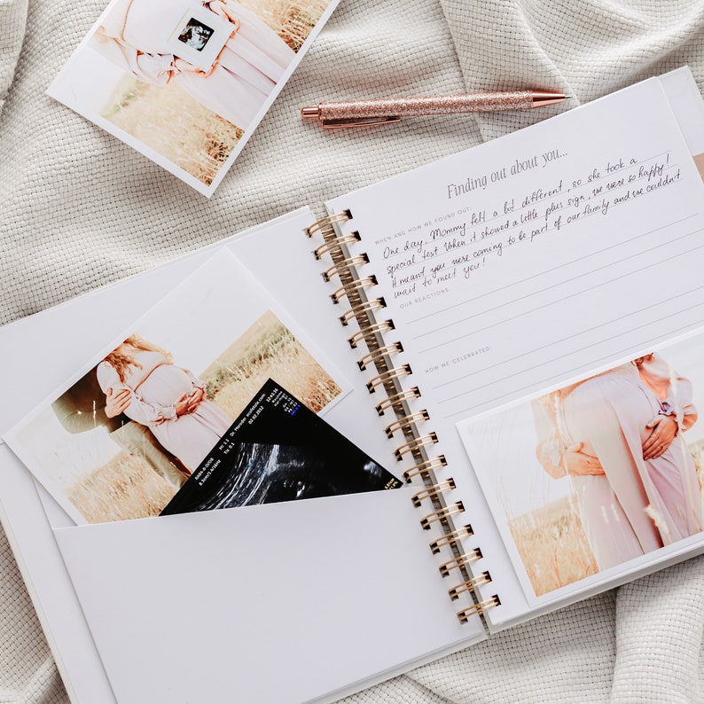 May include: A white spiral-bound journal with gold detailing, open to a page with handwritten text and a photo. Several photos and a pen with a glittery rose gold finish are also visible. The journal has pockets holding photos and an ultrasound image.
