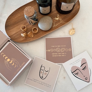 May include: A set of affirmation cards with pink and brown backgrounds. The cards feature gold lettering and illustrations of a face, a heart, and a crescent moon. The text on the cards includes phrases like "I am worthy of the very best in life" and "I trust the timing of my life".