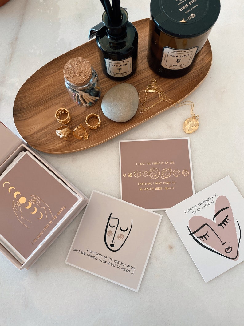 May include: A set of affirmation cards with pink and brown backgrounds. The cards feature gold lettering and illustrations of a face, a heart, and a crescent moon. The text on the cards includes phrases like "I am worthy of the very best in life" and "I trust the timing of my life".