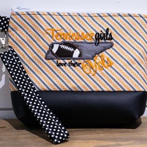 Op de afbeelding: Een zwarte en oranje gestreepte ritszak met een zwart-witte gestippelde band. De tas heeft een geborduurd ontwerp met een voetbal, de staat Tennessee en de tekst "Tennessee girls love their Vols".