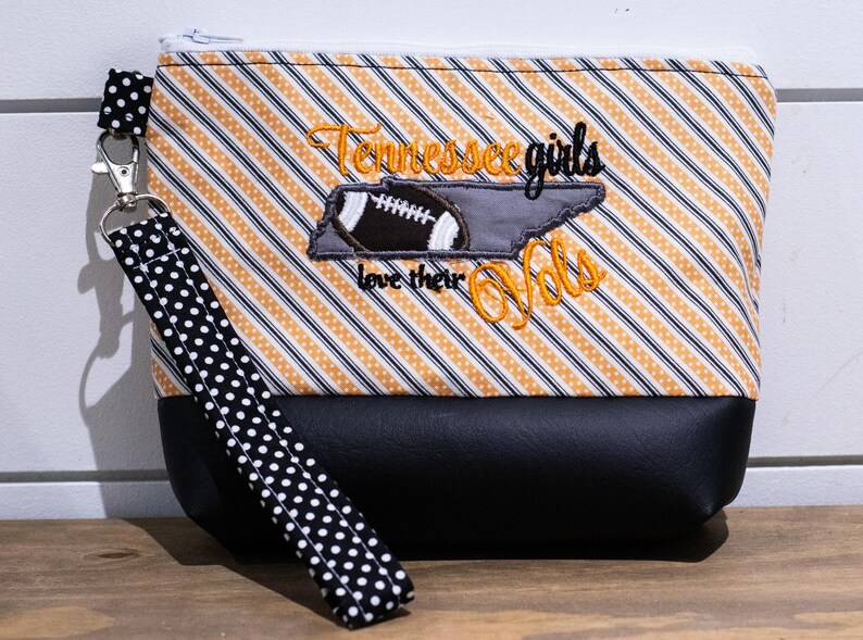Op de afbeelding: Een zwarte en oranje gestreepte ritszak met een zwart-witte gestippelde band. De tas heeft een geborduurd ontwerp met een voetbal, de staat Tennessee en de tekst "Tennessee girls love their Vols".