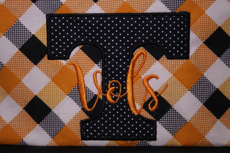 Op de afbeelding: Een zwart-wit geblokte stof met een zwarte vilt "T" met witte stippen en oranje borduurwerk dat "Vols" zegt.