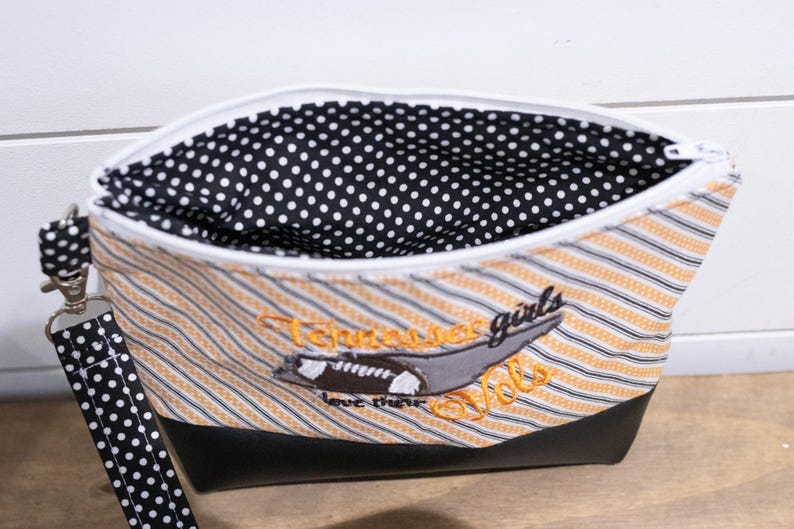 Op de afbeelding: Een zwarte en witte stippen-ritszak met een zwarte en oranje gestreepte buitenkant. De tas heeft een zwarte leren bodem en een zwarte en witte stippenband. De tas heeft een geborduurd ontwerp met een voetbal en de tekst "Tennessee girls love their Vols".