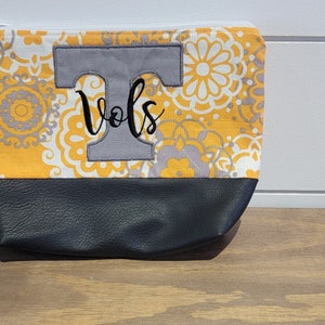Op de afbeelding: Een grijze en gele stoffen clutch met een grijze viltletter "T" en het woord "Vols" geborduurd op de voorkant. De clutch heeft een grijze rits en een grijze band.