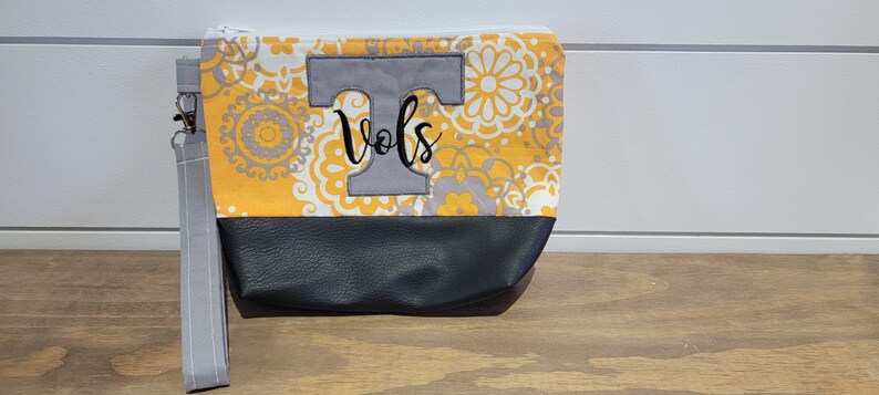 Op de afbeelding: Een grijze en gele stoffen clutch met een grijze viltletter "T" en het woord "Vols" geborduurd op de voorkant. De clutch heeft een grijze rits en een grijze band.