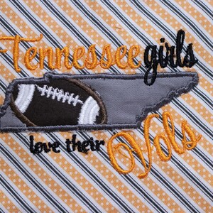 Op de afbeelding: Geborduurde patch met de tekst "Tennessee girls love their Vols" in oranje en zwarte draad. De patch heeft een grijze omtrek van de staat Tennessee met een bruine voetbal erin.