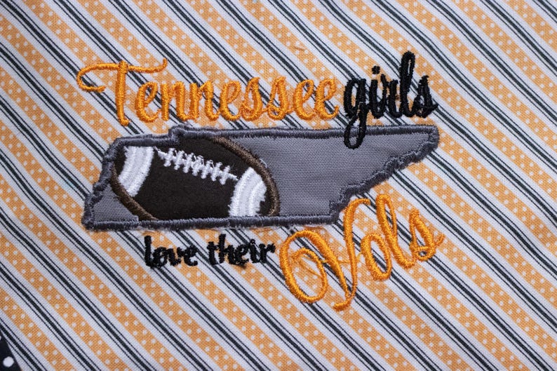 Op de afbeelding: Geborduurde patch met de tekst "Tennessee girls love their Vols" in oranje en zwarte draad. De patch heeft een grijze omtrek van de staat Tennessee met een bruine voetbal erin.