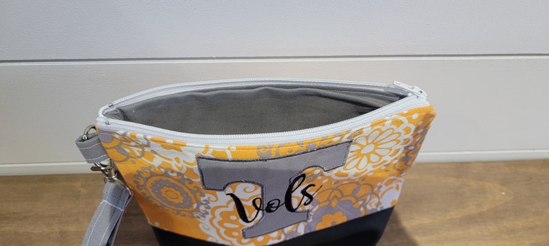 Op de afbeelding: Een gele en grijze ritszak met een wit bloemenpatroon. De tas heeft een grijze stoffen applicatie van een hoofdletter T met het woord "Vols" in zwart geborduurd.