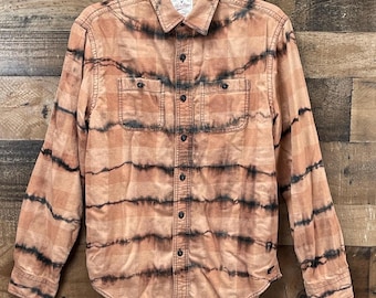 May include: A long-sleeve button-down shirt in a light orange and tan plaid pattern with horizontal black tie-dye stripes. The shirt features a pointed collar, a front button placket, and two chest pockets with buttoned flaps. The brand label reads American Eagle Outfitters.