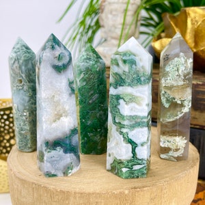 May include: Five moss agate crystal towers with varying patterns of green and white. The crystals are displayed on a round wooden base, showcasing their natural beauty and unique formations. The crystals are approximately 4-6 inches tall.