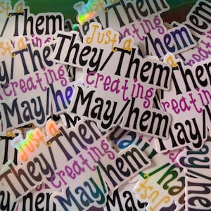 May include: A close-up of several stickers with the text "Just a They/Them Creating May/Them" in black and purple lettering on a white background.