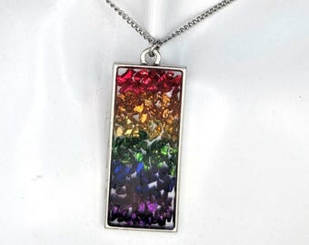 May include: A rectangular pendant necklace featuring a silver-toned metal frame filled with small, crushed stone fragments arranged in a vertical rainbow gradient of red, orange, yellow, green, blue, and purple, suspended on a silver-toned chain.