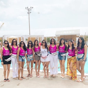 May include: A group of women wearing pink tank tops with the text "the party" and white fanny packs. The woman in the centre is wearing a white tank top with the text "the wife". They are standing in front of a swimming pool.