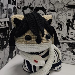 May include: A handmade crochet doll with a tan face, black hair, and large, round eyes. The doll wears a white and navy striped outfit and has a white snake wrapped around its neck. The doll is inspired by anime.