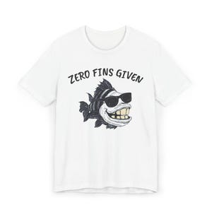 May include: White t-shirt featuring a cartoon fish wearing sunglasses and a wide grin. The fish is black and white with a striped pattern. The text "ZERO FINS GIVEN" is printed above the fish in black, bold letters.