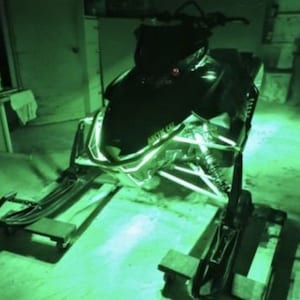 May include: A black snowmobile with green LED lights under the chassis. The snowmobile is on a black metal dolly.