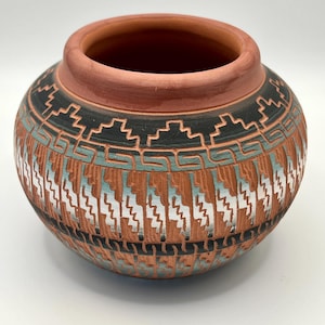 May include: A handcrafted, round terracotta pot with a wide opening. The pot features a detailed geometric pattern in black, turquoise, and white against a reddish-brown background. The design includes stepped and zigzag motifs, showcasing traditional craftsmanship.