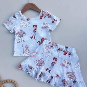 May include: A white two-piece outfit featuring a short-sleeved top and a skirt. The set is adorned with a repeating pattern of cartoon characters in various poses. The skirt has a ruffled hem and a drawstring waist.